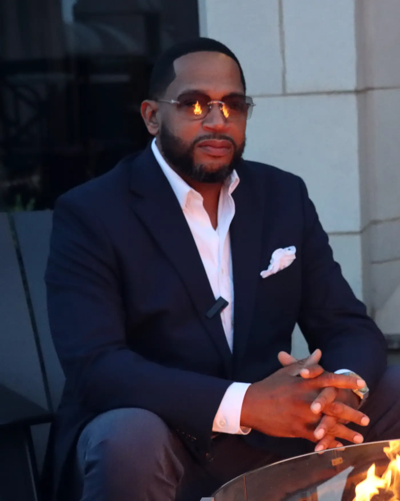 Kevin Maples seated beside a lit fire pit at dusk, hands clasped, in a navy suit and open white shirt